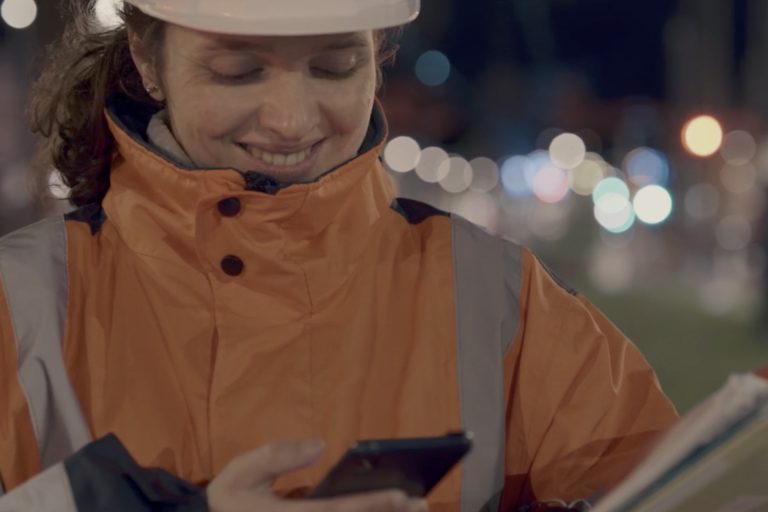 Femme portant un casque et une combinaison de chantier qui regarde son téléphone portable en souriant